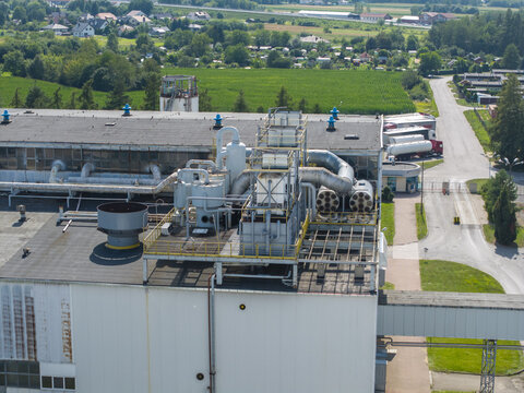 Aerial view of sugar refinery technical installations with industrial pipes and equipment on the rooftop, surrounded by green fields and a road with trucks in Poland.