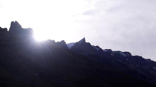 Dramatic cinematic timelapse of bright sunlight and moving clouds behind a dark, jagged, silhouetted mountain ridge in the Himalayas.