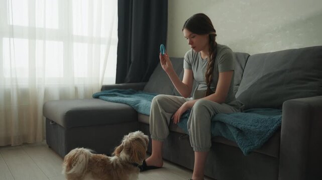 Woman checking phone while dog watches from floor, subdued natural light through large window, casual seated pose on sofa with braided hair, understated modern interior, gentle domestic pause blending