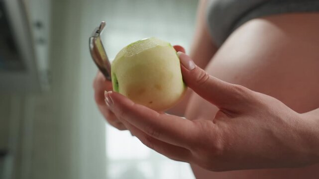 Pregnant white belly closeup peeling apple with careful hands and peeler near exposed bump, soft window light, tactile peel falling, focus on prenatal nutrition, texture and grip, mindful kitchen