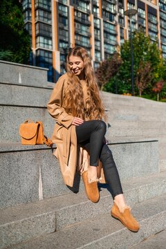 Young smiling woman in tan trench coat seated on stone steps in urban cityscape with beige handbag and modern buildings behind, stylish outdoor portrait, with woman, female, adult, smile detail