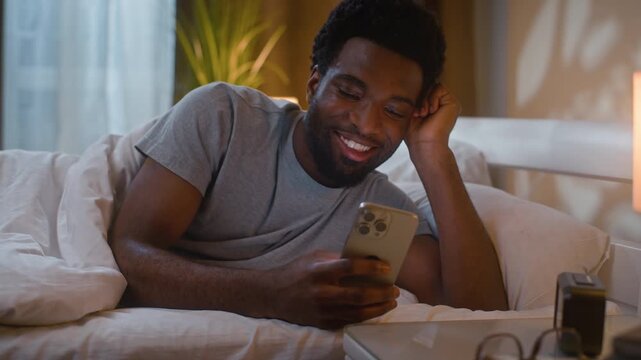Happy African American man laughing using mobile phone in bedroom evening scrolling social media watching funny video smartphone sleep ethnic guy male relaxing at home bed night cheerful client user
