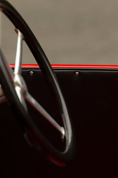 Tight composition showing a glossy black steering wheel with metal spokes in the foreground and a textured black door panel with red trim along the top edge