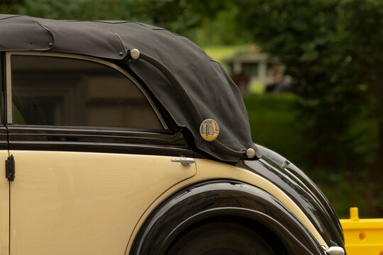 Close-up of the rear side of a cream and black vintage convertible showing the folded black soft top, rounded rear fender, chrome trim, and side mirror. The