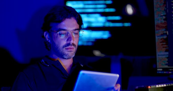 Adult Asian male cybersecurity analyst reviews tablet, then looks toward code monitors while checking information during network security monitoring, system analysis, and secure data operations.
