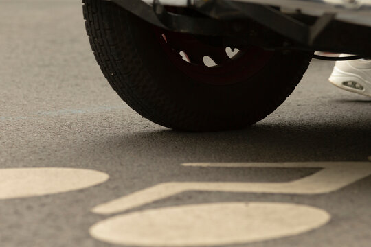 Low-angle view showing the rear wheel of a vehicle hovering above marked asphalt with large painted symbols and a pedestrian's white shoe visible at the edge