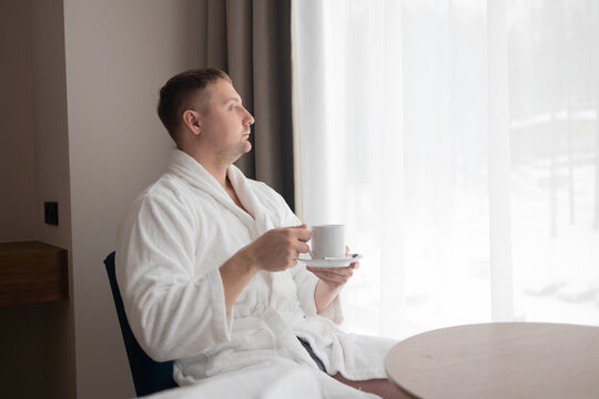 Side view profile of a relaxed millennial male holding a cup of tea while sitting in an armchair near a large window. Concept of morning routine, relaxation, luxury spa wellness, and hospitality
