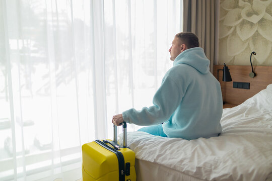 Side view of a male traveler holding luggage handle while resting on a couch or bed near window. Concept of travel arrival, hotel check-in, cozy tourism accommodation, and departure waiting.