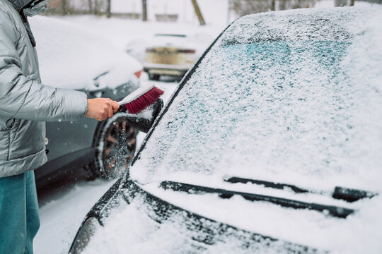 Close-up of a person clearing fresh white snow off an automobile front window and wipers after blizzard. Winter vehicle maintenance, automotive care, and freezing weather concept.