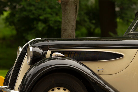 Close-up view of the front left fender, headlight, wheel and decorative grille of a classic black and cream vintage automobile parked outdoors with soft