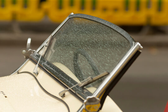 Detailed close-up of a cream-colored vintage car windshield covered in raindrops, framed by polished chrome windshield frame and dual wipers. Soft natural