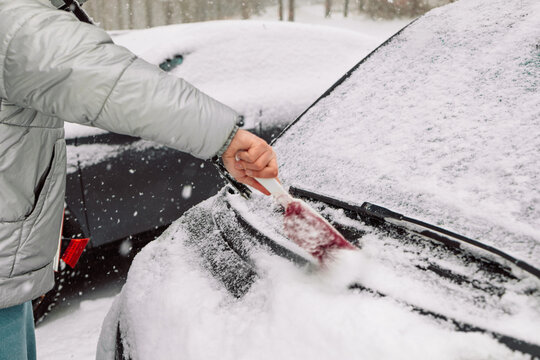 Close-up of a person clearing fresh white snow off an automobile front window and wipers after blizzard. Winter vehicle maintenance, automotive care, and freezing weather concept.