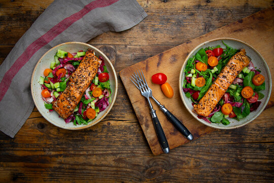 Two bowls of grilled salmon salad sit on a dark wooden table