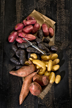 Different sorts of potatoes and peeler on brown paper, jute and dark wood
