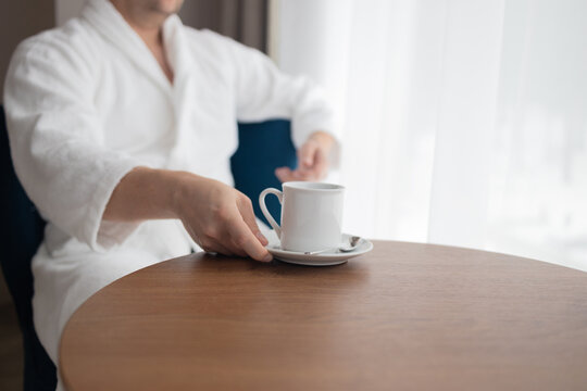 Side view profile of a relaxed millennial male holding a cup of tea while sitting in an armchair near a large window. Concept of morning routine, relaxation, luxury spa wellness, and hospitality