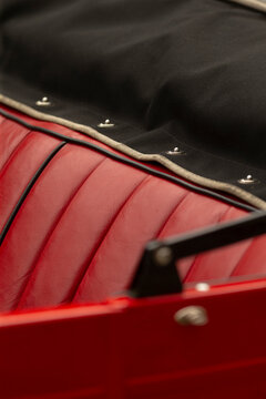 Close-up view of a red leather car seat with vertical stitching beside the folded black convertible top edge. Visible chrome snaps and beige piping attach the