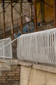 Close-up view of a curved white metal railing mounted on a concrete wall in an aged outdoor seating or walkway area. The scene shows tubular vertical bars