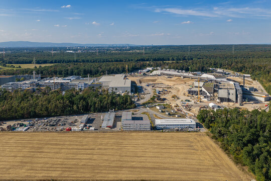 Aerial view of the Facility for Antiproton and Ion Research construction site with modern laboratory buildings, heavy machinery, and surrounding forest in Darmstadt, Hessen, Germany.