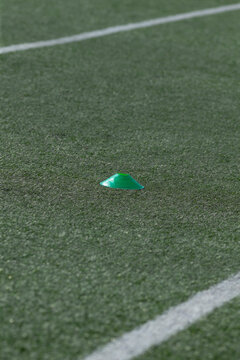 A small green training cone placed on textured artificial turf beside a white painted field line. The scene shows close-up surface detail of the synthetic