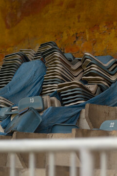 Disused light blue plastic stadium seats piled in rows and stacks, some bearing white seat numbers, partially draped with a blue tarpaulin. The seating