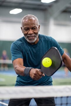 Smiling mature man playing intense pickleball game with bright yellow ball inside modern indoor sports facility