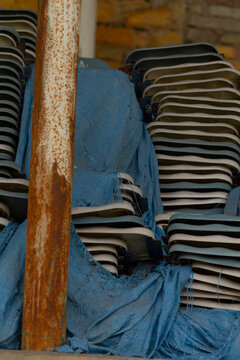 A vertical composition showing folded plastic chairs stacked tightly against a brick wall, partially covered by a faded blue tarp. A rusted metal post stands