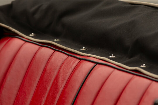 Tight detail of a red leather car bench seat showing vertical stitched pleats and black piping, topped by the edge of a black convertible top or protective
