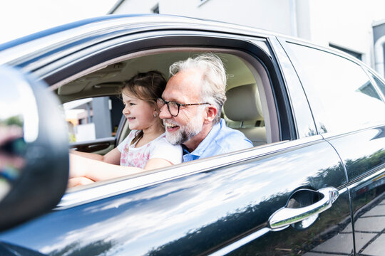 Little girl sitting on lap of grandfather, pretending to steer the car