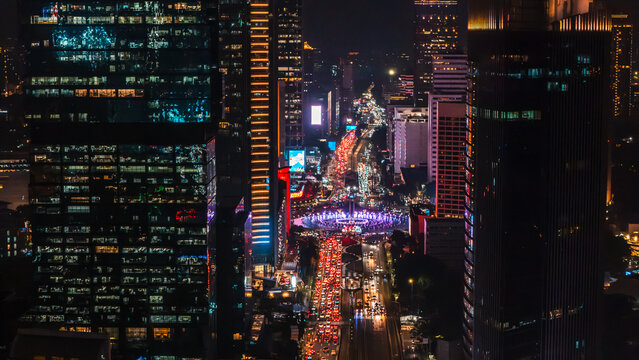 Aerial view of the illuminated Bundaran HI roundabout at night, surrounded by towering skyscrapers and heavy traffic flow in Jakarta, Indonesia.