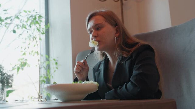 White woman eating salad by window, natural light, indoor cafe, business blazer, thoughtful pause while tasting, pensive expression, plants visible through glass, minimalist bowl and fork, calm midday
