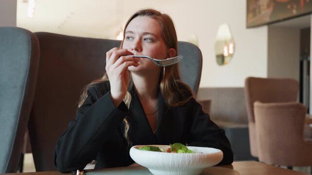 Businesswoman enjoying salad in cafe, poised woman in blazer seated in armchair, white bowl of greens, thoughtful gaze between bites, bright bistro interior, natural light, upscale relaxed atmosphere