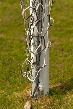 Vertical close-up view of a white soccer goalpost with a tangled nylon goal net draped and looped around the post, set on a sunlit grassy field. The image
