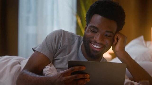 Laughing relaxed African American man holding tablet device lying on bed homeowner client male guy watching funny movie scrolling social media video film comedy online late night home bedroom indoors