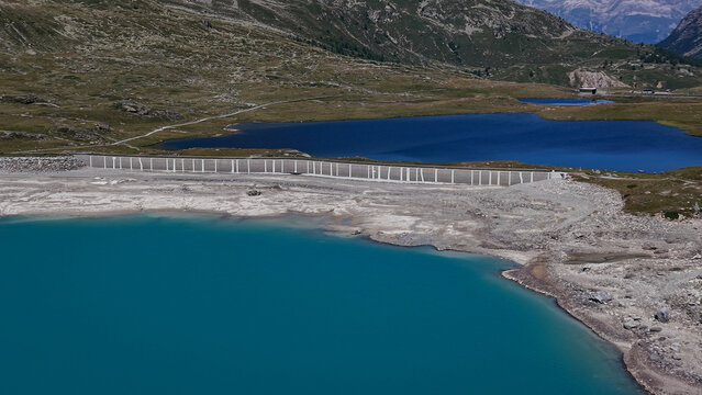 Aerial view of Lago Bianco reservoir with turquoise water and concrete dam, surrounded by the rugged alpine landscape of the Bernina Pass in Poschiavo, Grisons, Switzerland.