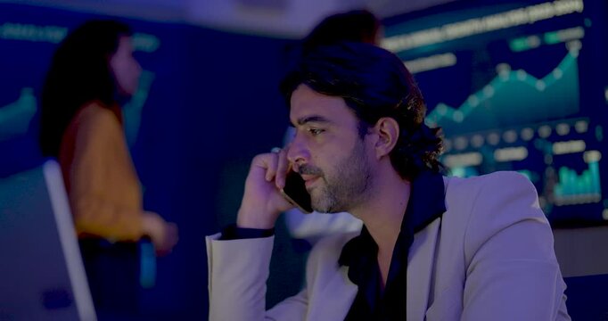 Adult male professional talks on smartphone while two female colleagues discuss digital performance dashboard behind him, combining corporate communication with collaborative business data analysis.