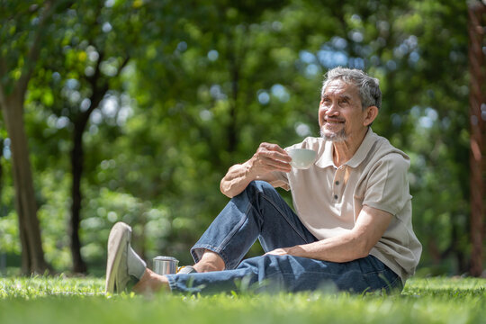 happy asian senior man enjoying peaceful retirement lifestyle holding a teacup,sitting and relaxing on the lawn in nature park