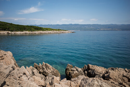 Sharp limestone rocks in the foreground along the pristine coastline of Krk Island, Croatia, overlooking clear turquoise Adriatic waters, a green pine-covered peninsula under a bright blue sky.