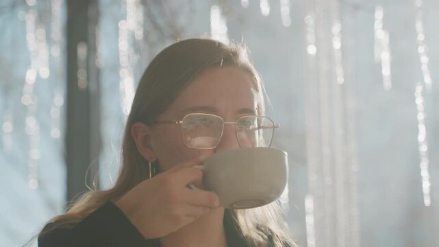 Woman tasting coffee with surprised wince, bright backlight from icicle laced window, evaluates bitter flavor, adjusts glasses, candid cafe scene of tasting critique by freelance writer
