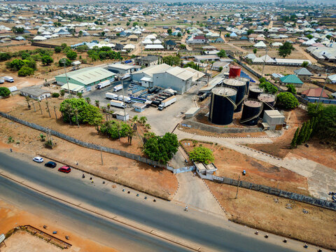 Aerial view of the Indomie Factory with industrial warehouses, large storage tanks, and parked trucks, surrounded by residential houses and dry landscape in Kura, Kaduna, Nigeria.
