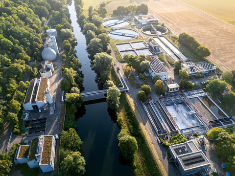 Aerial view of a wastewater treatment plant situated beside a river, surrounded by lush trees and agricultural fields in Germany.