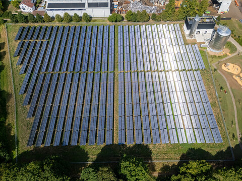 Aerial view of a solar thermal energy farm with rows of panels reflecting sunlight, surrounded by green grass and trees in Germany.