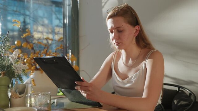 Sunlit table scene caucasian designer reviewing samples and clipboard notes, comparing swatches and colors beside vase and glass, concentrated expression and deliberate gestures, studio planning