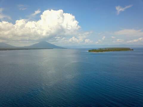 Aerial view of a tropical sea featuring a small island and a distant volcano under a blue sky with white clouds in Indonesia.