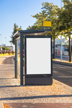Modern urban bus stop with a blank advertising display in Lisbon, Portugal. The shelter features a contemporary dark frame, glass panels and a yellow bus stop sign, surrounded by trees and Portuguese 
