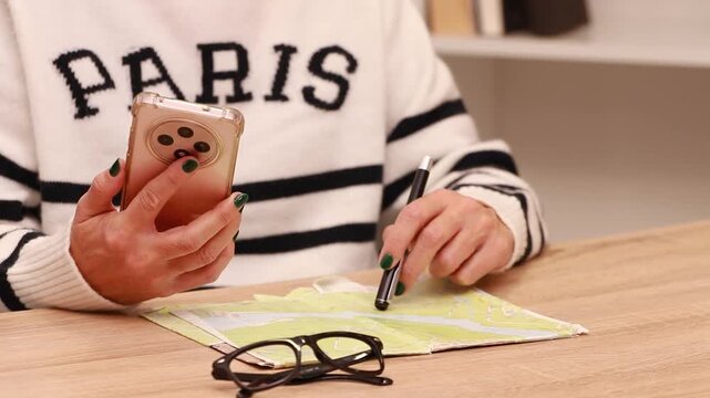 young woman's hands with a mobile phone reviewing the trip or itinerary on the map on the desk, wearing a Paris sweater