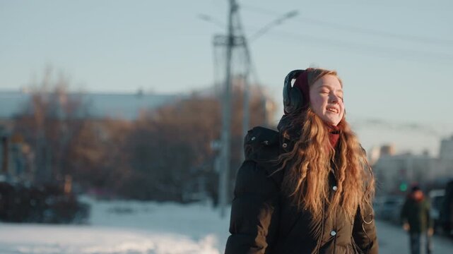 White woman smiling headphones winter city portrait, radiant grin with red gloves and long curly hair, black puffer coat and warm low sun on face, pedestrians and cars blurred in background, upbeat