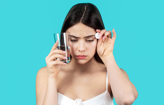 Woman take some pills, holds glass of water, isolated on blue. Young woman taking pill against headache. Female taking a pill with a glass of water. Woman taking drugs to relieve headache
