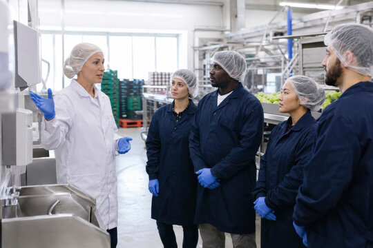 Supervisor gives safety instructions to factory workers wearing hairnets and gloves in a food production facility. The team listens attentively near processing equipment.