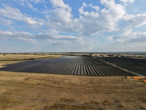 Aerial view of a large solar farm with rows of photovoltaic panels set in a rural landscape under a blue sky in Irchester, England, United Kingdom.