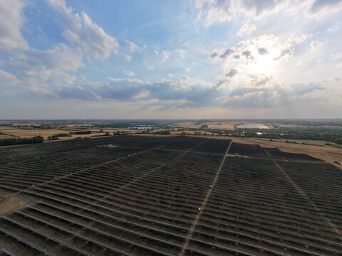 Aerial view of a large solar farm with rows of photovoltaic panels in a rural landscape under a bright sky with sunbeams in Irchester, England, United Kingdom.
